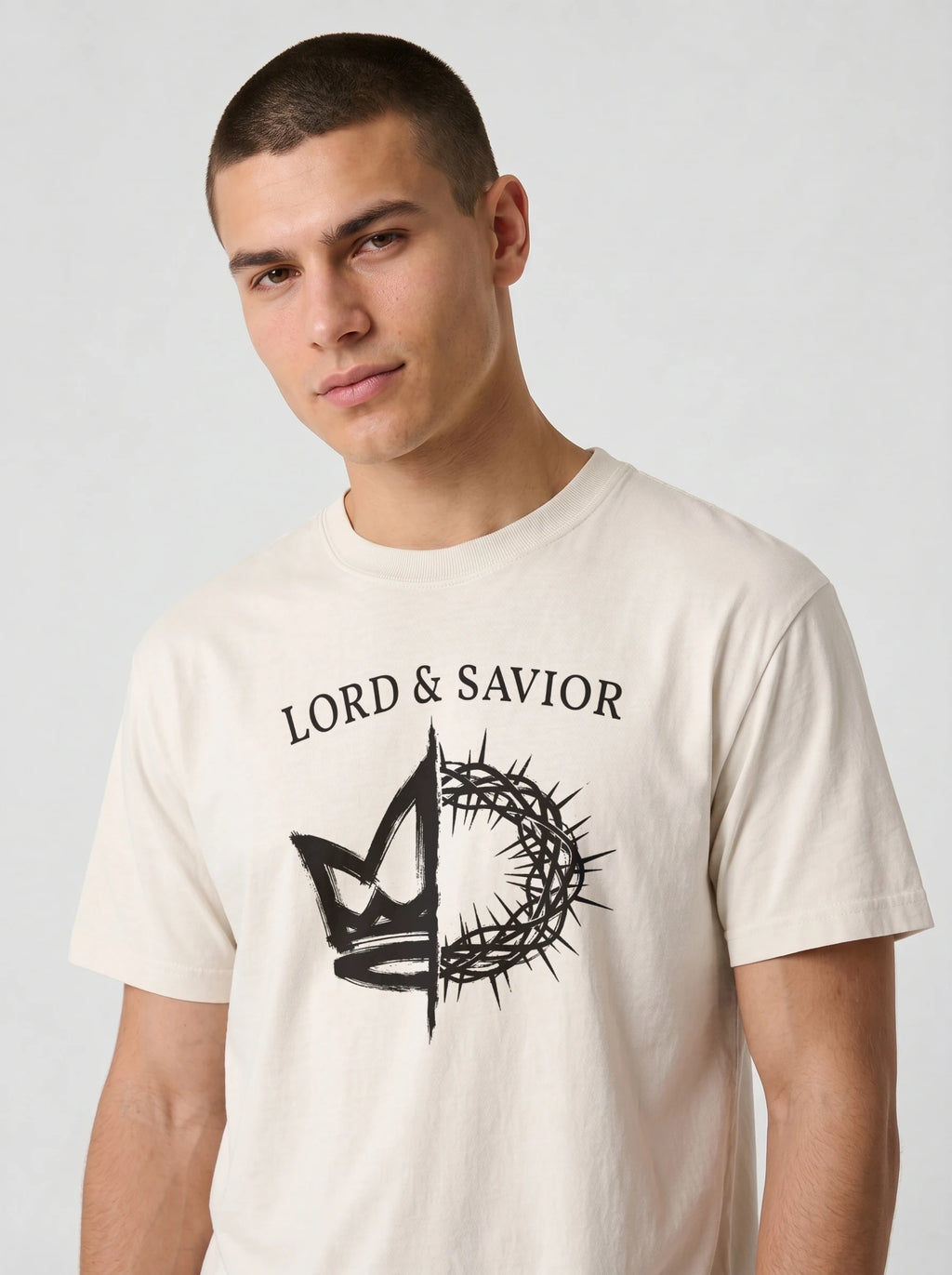 A man wearing a cream-colored t-shirt with a black graphic print that reads 'LORD & SAVIOR' above a crown and thorn design, photographed against a light gray background.