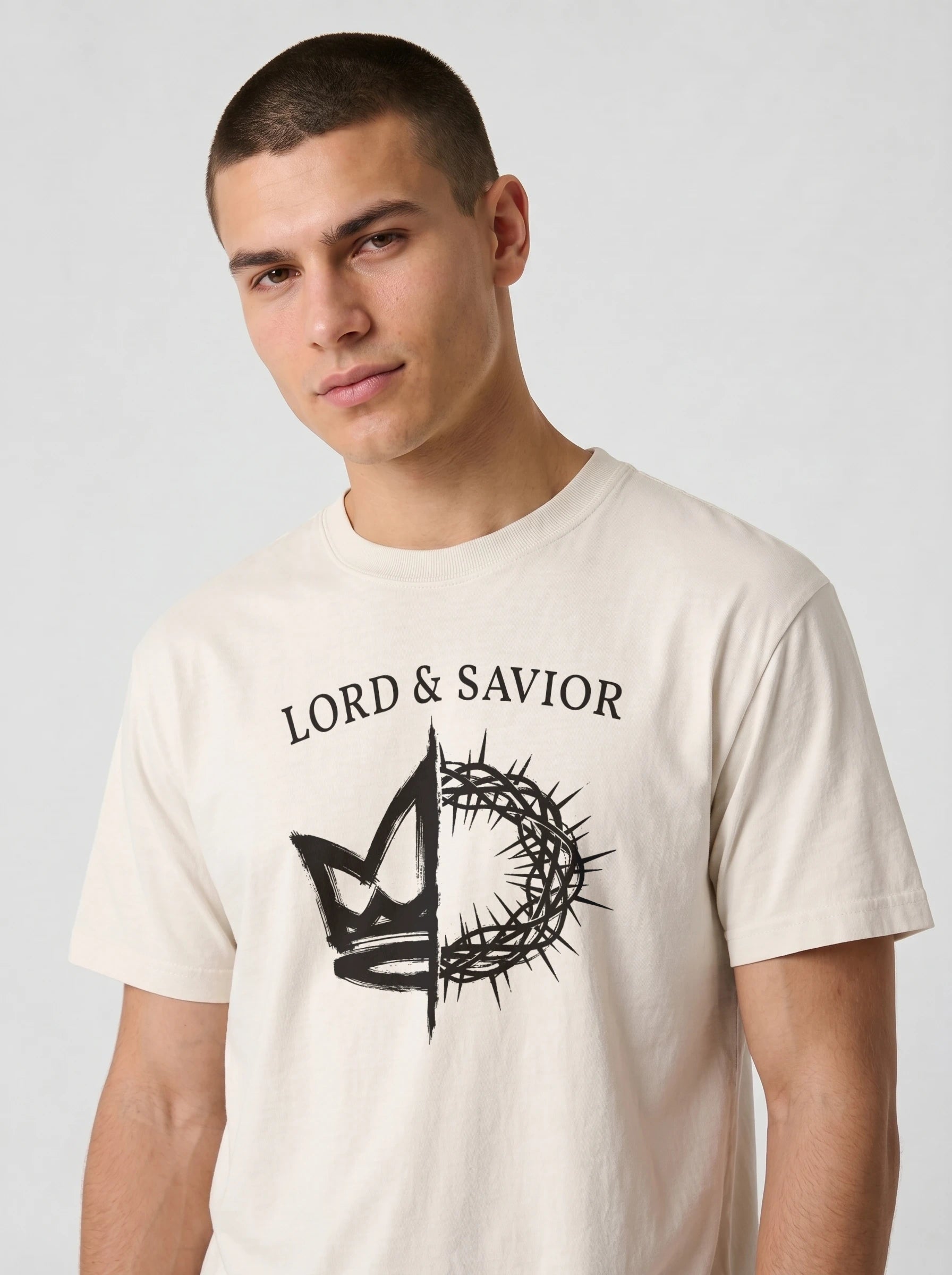 A man wearing a cream-colored t-shirt with a black graphic print that reads 'LORD & SAVIOR' above a crown and thorn design, photographed against a light gray background.