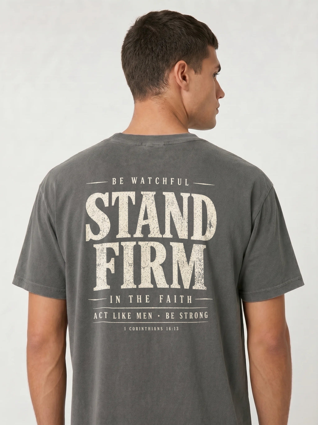 A man wearing a dark gray t-shirt, viewed from the back. The shirt features a large distressed white graphic print on the back with the words 'STAND FIRM' and smaller text above and below.