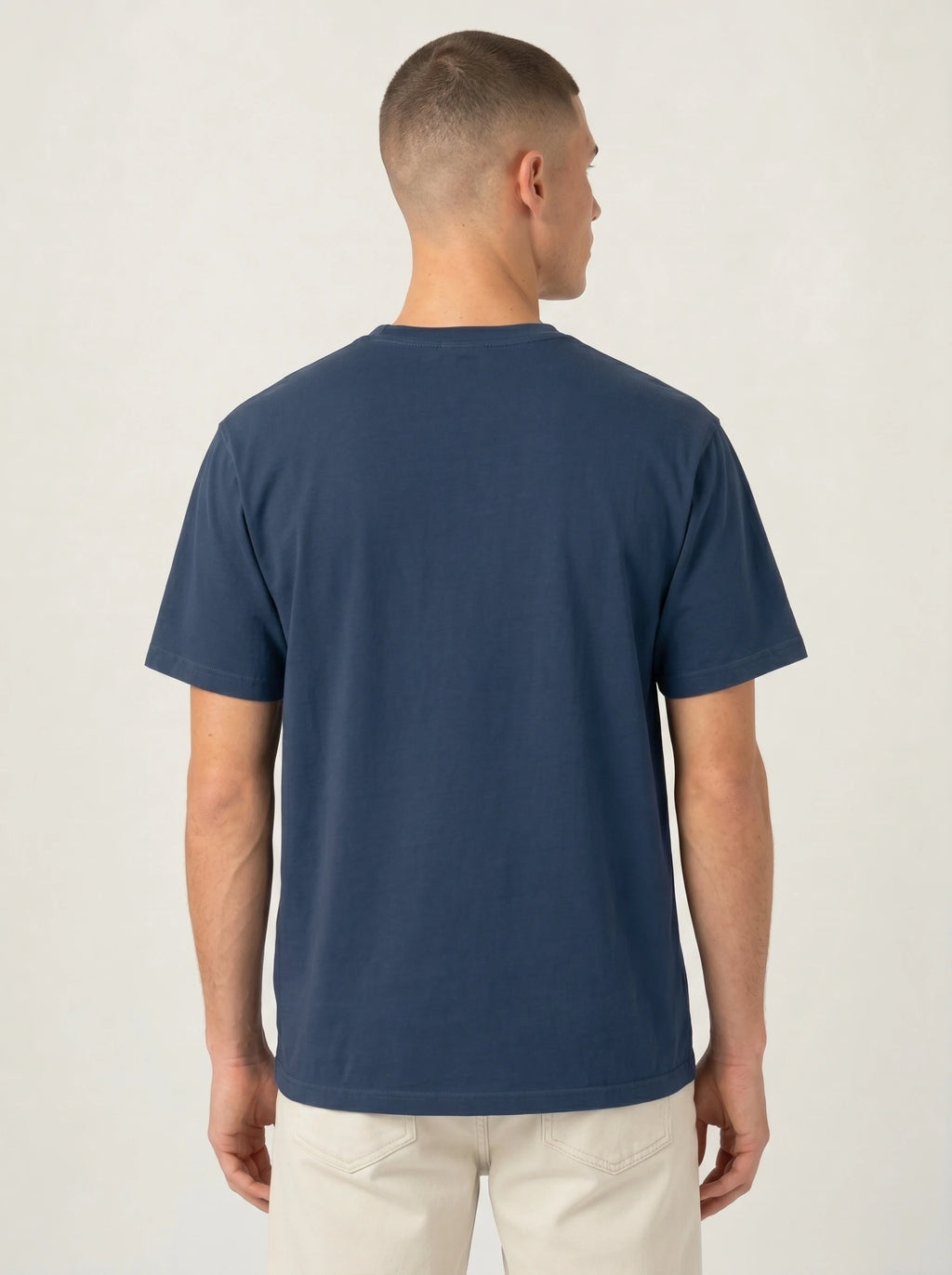 A man viewed from the back wearing a solid navy blue short-sleeve crewneck t-shirt and off-white trousers, set against a plain light gray studio background.