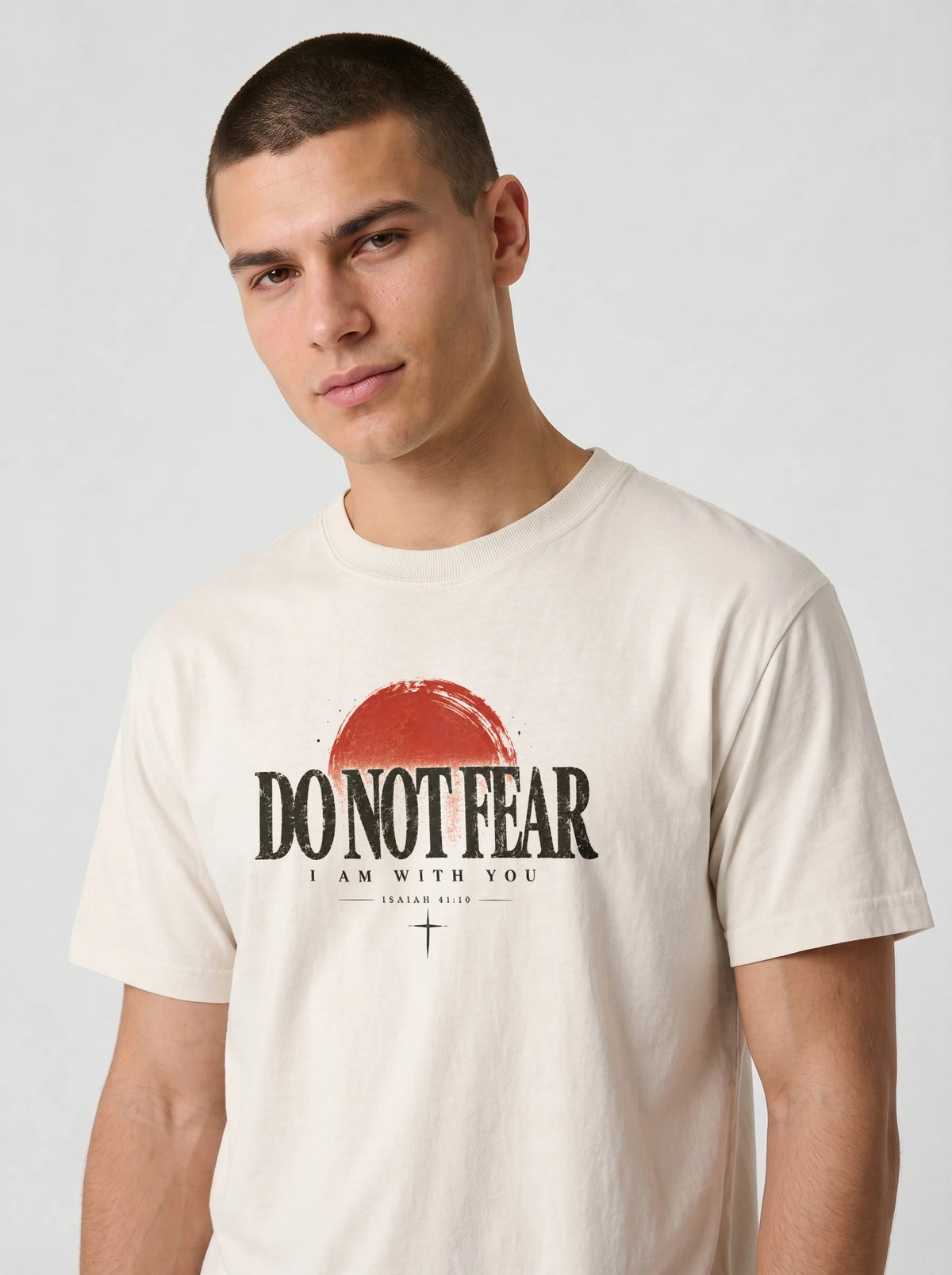 A man wearing a cream-colored t-shirt with a graphic print that reads 'DO NOT FEAR I AM WITH YOU' under a red sun graphic, on a light gray background.