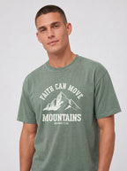 A man wearing a muted sage green graphic t-shirt with a white mountain design and the text 'FAITH CAN MOVE MOUNTAINS' on the chest, set against a plain light gray studio background.