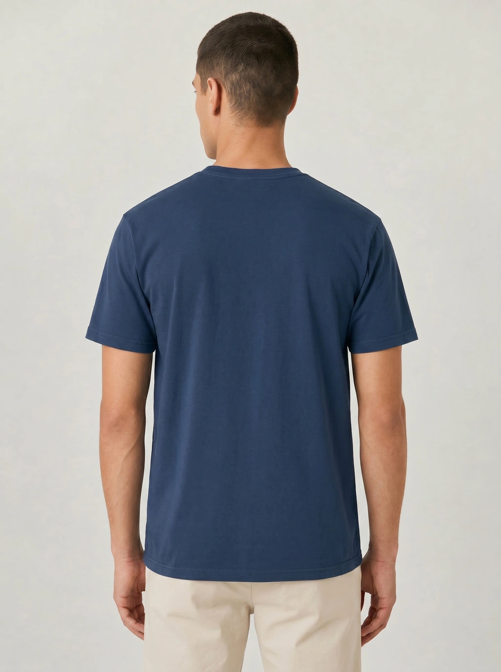 A man viewed from the back wearing a solid navy blue short-sleeve crewneck t-shirt and light beige trousers, set against a plain light gray studio background.