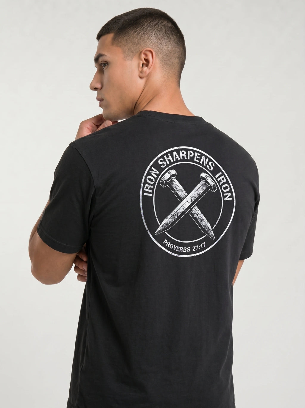 A man wearing a black t-shirt, viewed from the back. The shirt features a large white circular graphic on the back with the text 'IRON SHARPENS IRON' and 'PROVERBS 27:17' surrounding an illustration of two crossed nails.