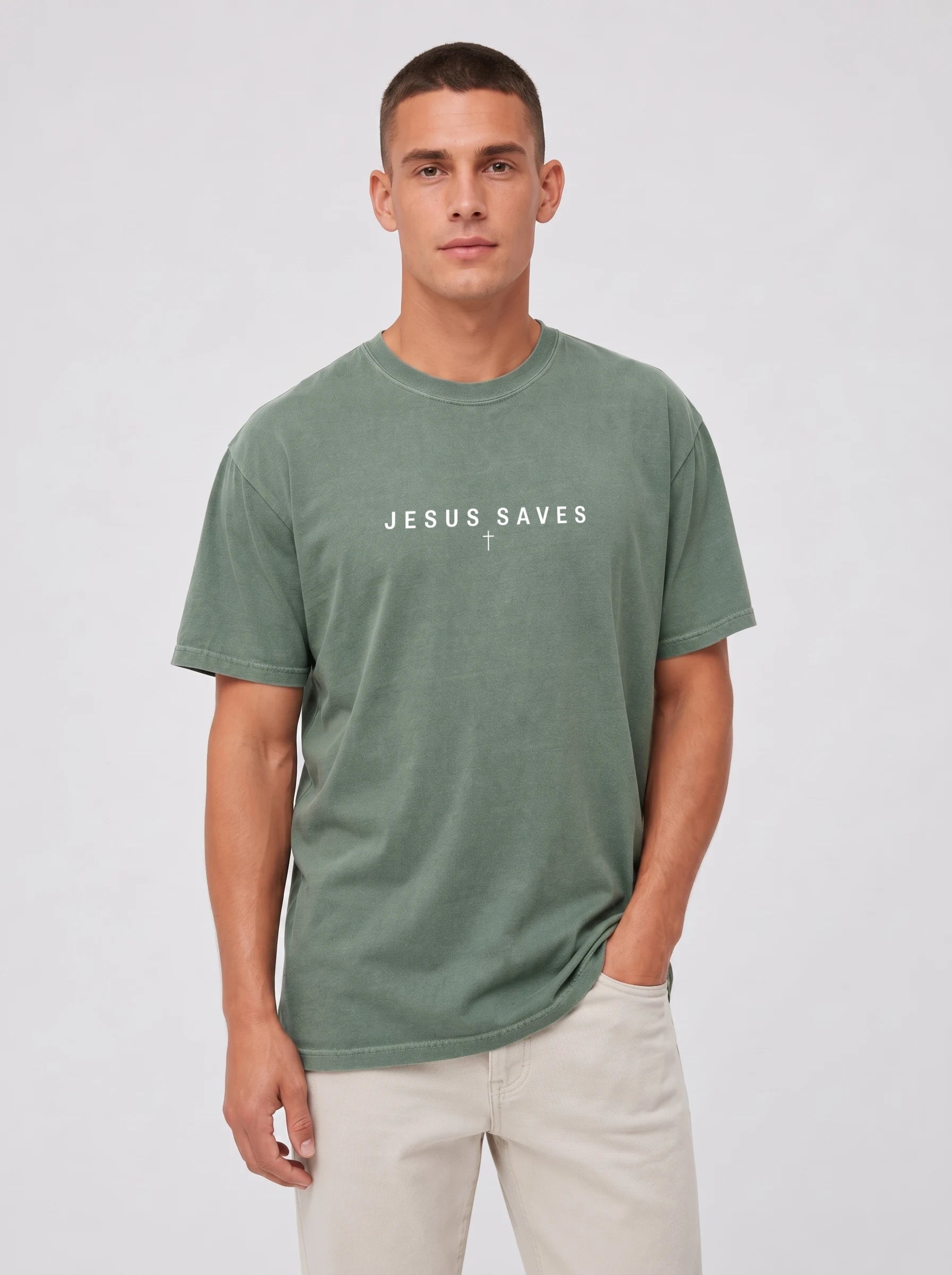 A man wearing a muted sage green t-shirt with the text 'JESUS SAVES' printed in white across the chest, paired with light beige pants, against a light gray studio background.