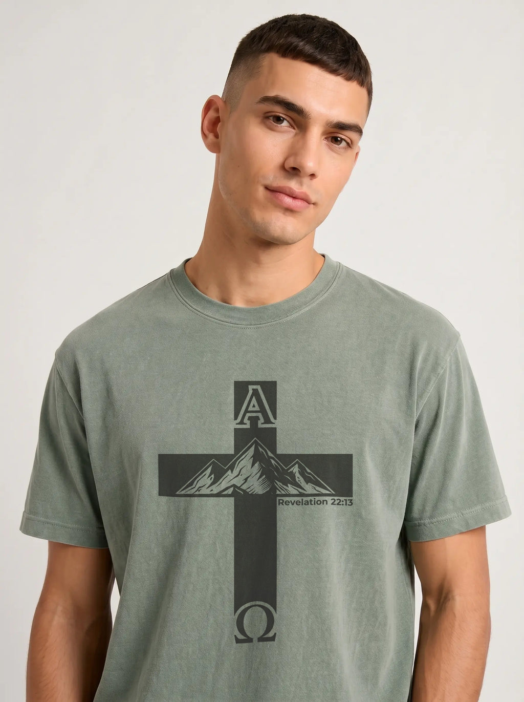 A man wearing a mineral-washed sage green t-shirt with a black graphic of a cross, mountains, and the Greek letters Alpha and Omega with 'Revelation 22:13' printed on the front, against a light gray background.