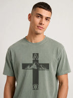 A man wearing a mineral-washed sage green t-shirt with a black graphic of a cross, mountains, and the Greek letters Alpha and Omega with 'Revelation 22:13' printed on the front, against a light gray background.