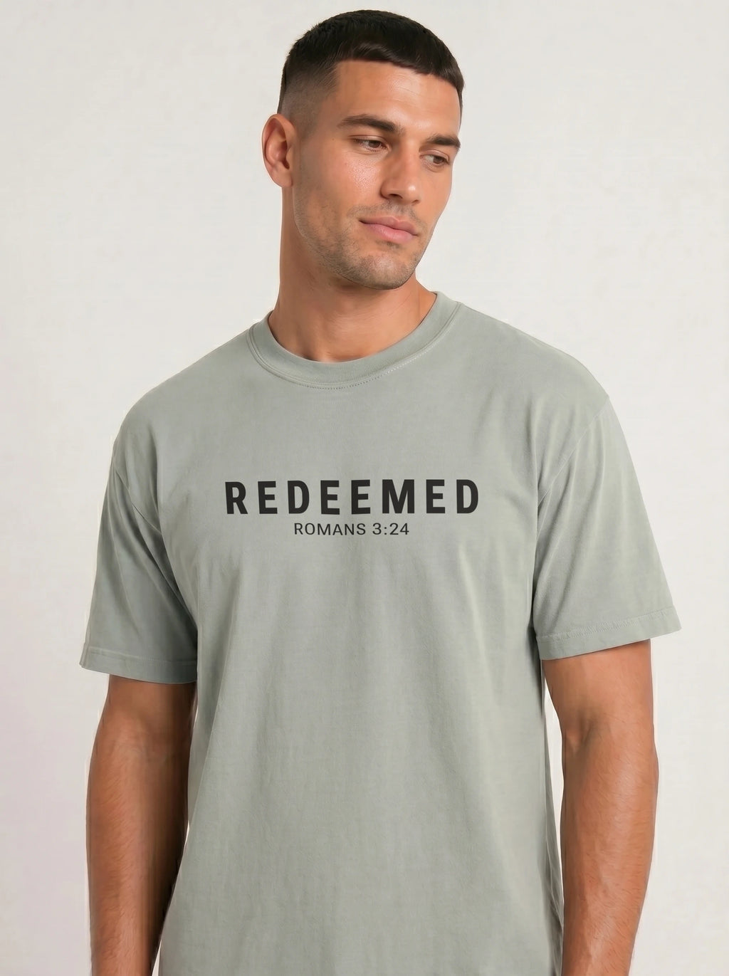 A man wearing a light sage green t-shirt with the text 'REDEEMED ROMANS 3:24' printed in black across the chest, set against a plain light gray background.