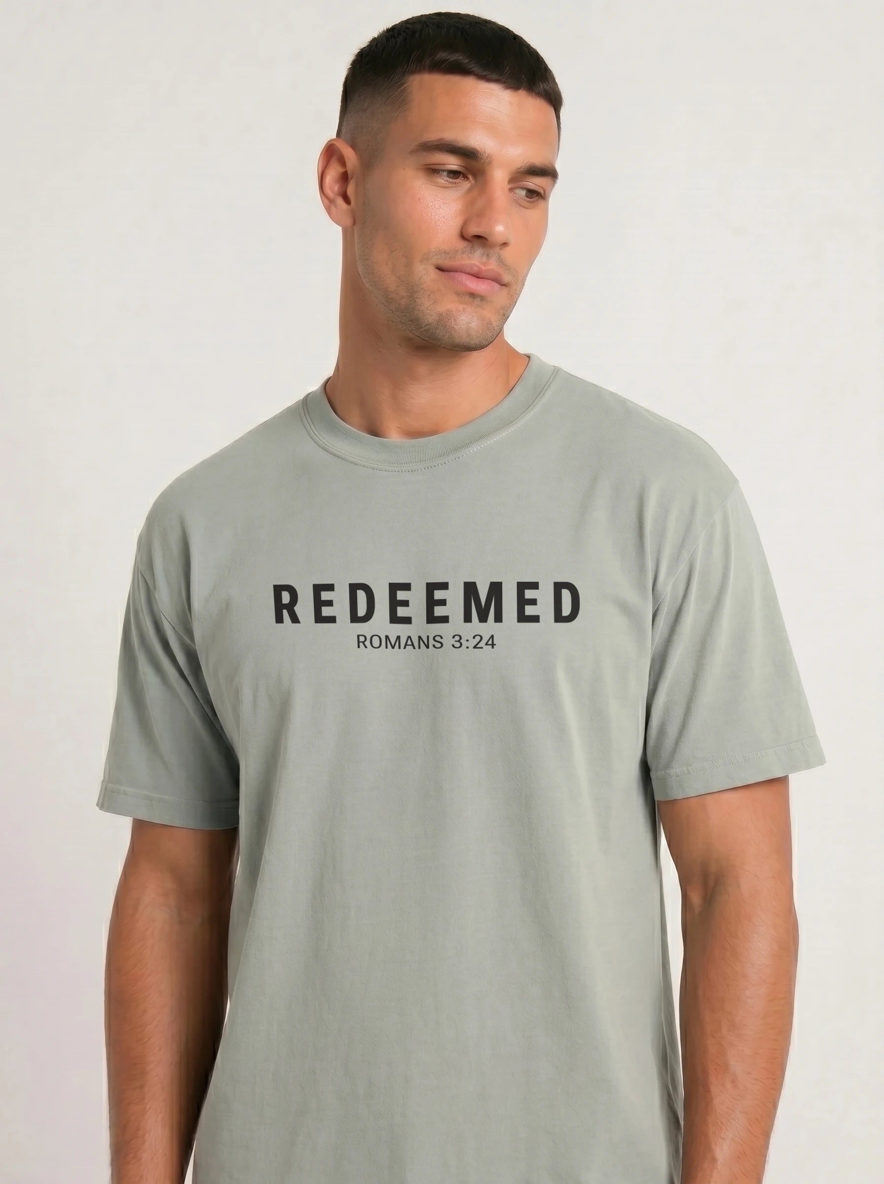 A man wearing a light sage green t-shirt with the text 'REDEEMED ROMANS 3:24' printed in black across the chest, set against a plain light gray background.