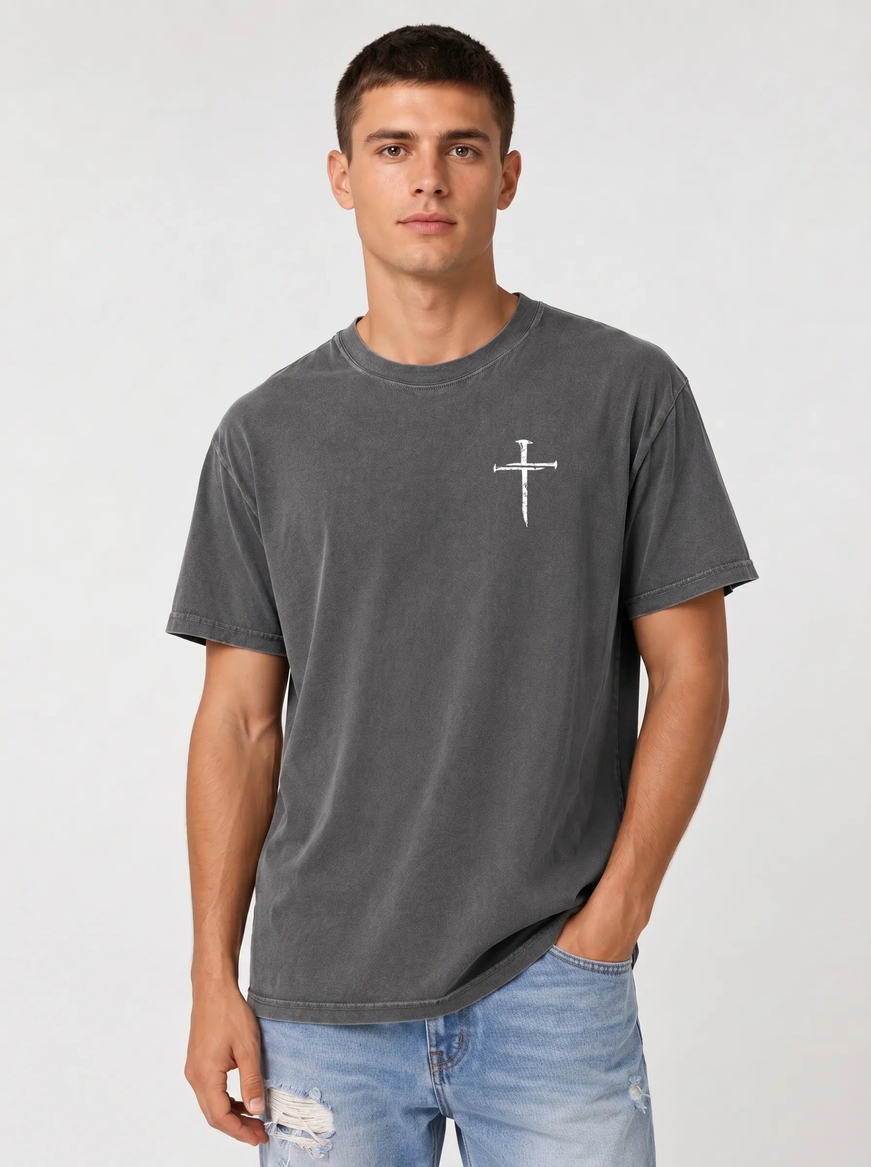 A man wearing a dark gray mineral-wash t-shirt with a small white cross graphic on the chest, paired with light blue jeans, against a light gray studio background.