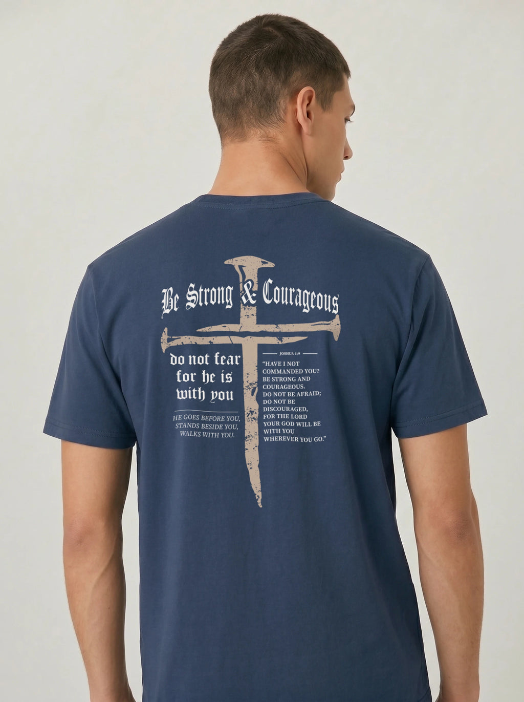 A man wearing a navy blue t-shirt, viewed from the back. The shirt features a large graphic print of a cross , accompanied by the words 'Be Strong & Courageous' and a Bible verse.