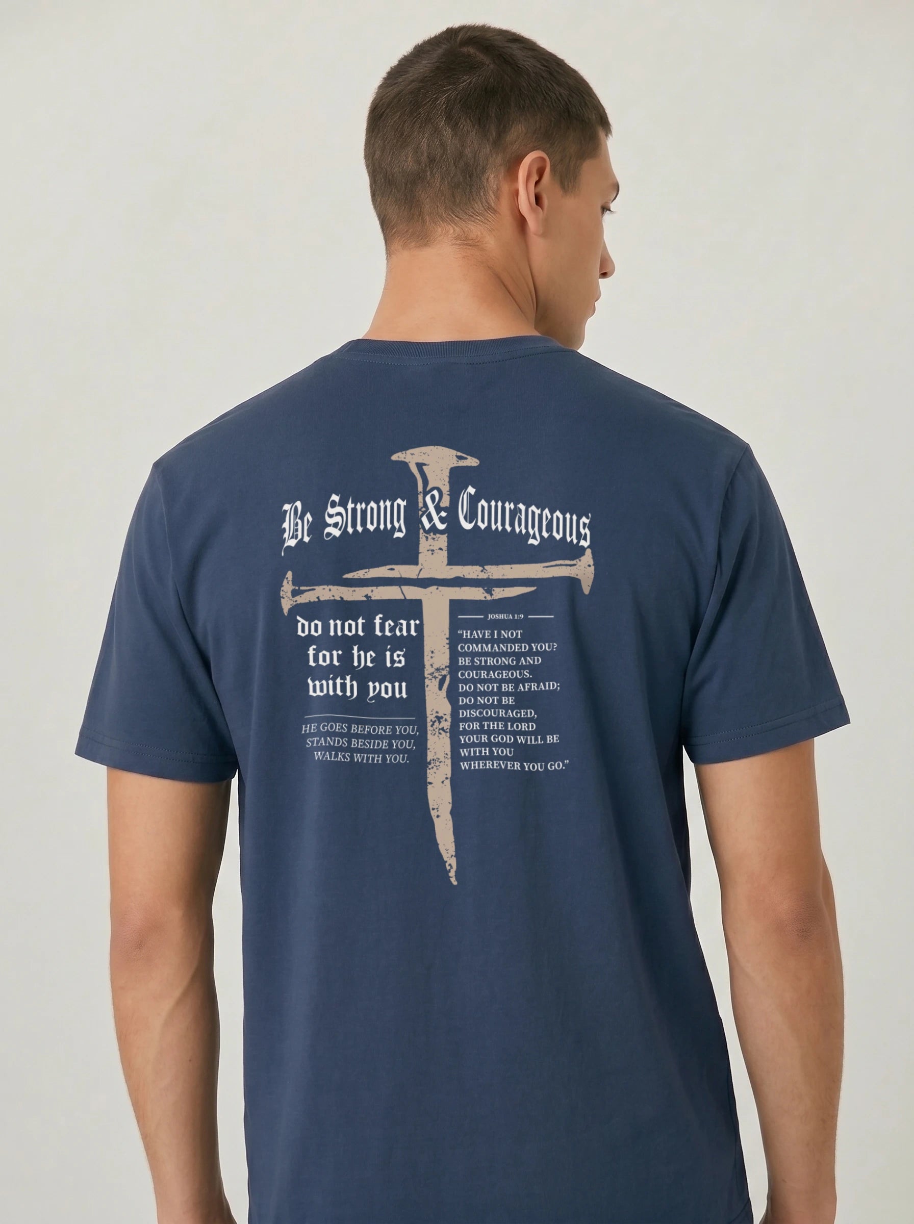 A man wearing a navy blue t-shirt, viewed from the back. The shirt features a large graphic print of a cross , accompanied by the words 'Be Strong & Courageous' and a Bible verse.