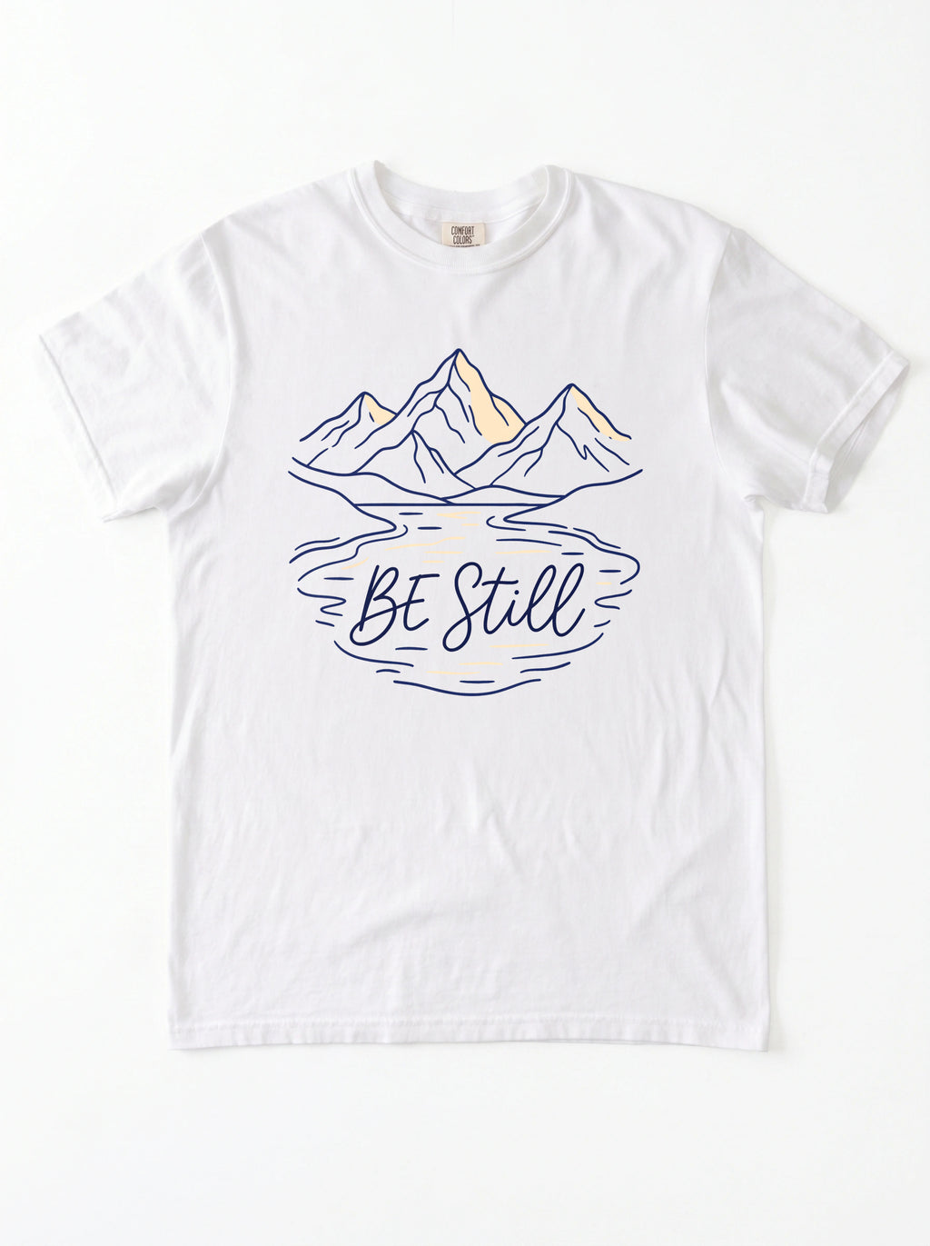 A white short-sleeve crewneck t-shirt featuring a graphic of mountains and a lake reflection with the text 'Be Still' in navy blue and light yellow accents.