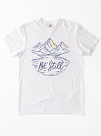 A white short-sleeve crewneck t-shirt featuring a graphic of mountains and a lake reflection with the text 'Be Still' in navy blue and light yellow accents.