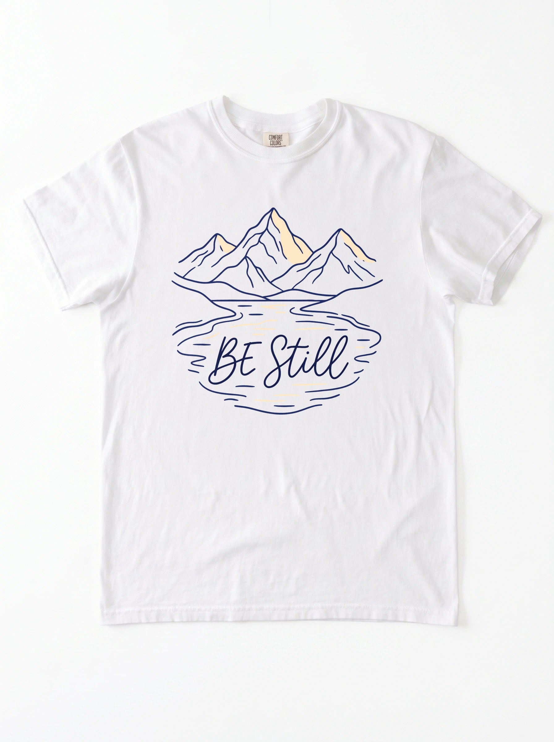 A white short-sleeve crewneck t-shirt featuring a graphic of mountains and a lake reflection with the text 'Be Still' in navy blue and light yellow accents.