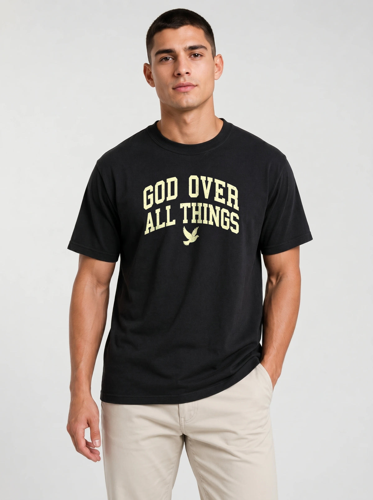 A man wearing a black short-sleeve t-shirt with 'GOD OVER ALL THINGS' printed in a distressed cream font on the chest, paired with light beige pants, against a light gray studio background.