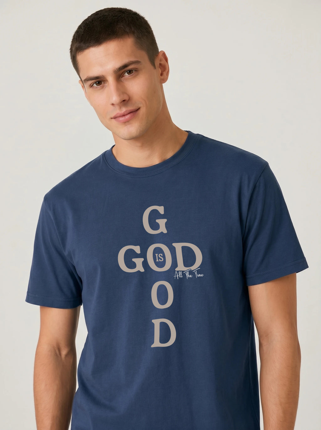 A man wearing a navy blue crewneck t-shirt with a vertical graphic text design that reads 'GOD IS ALL THE TIME' in beige, set against a plain light gray background.