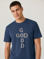 A man wearing a navy blue crewneck t-shirt with a vertical graphic text design that reads 'GOD IS ALL THE TIME' in beige, set against a plain light gray background.