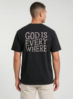 A man viewed from the back wearing a black t-shirt with the phrase 'GOD IS EVERY WHERE' printed in distressed white capital letters across the back, paired with light beige pants, against a light gray studio background.