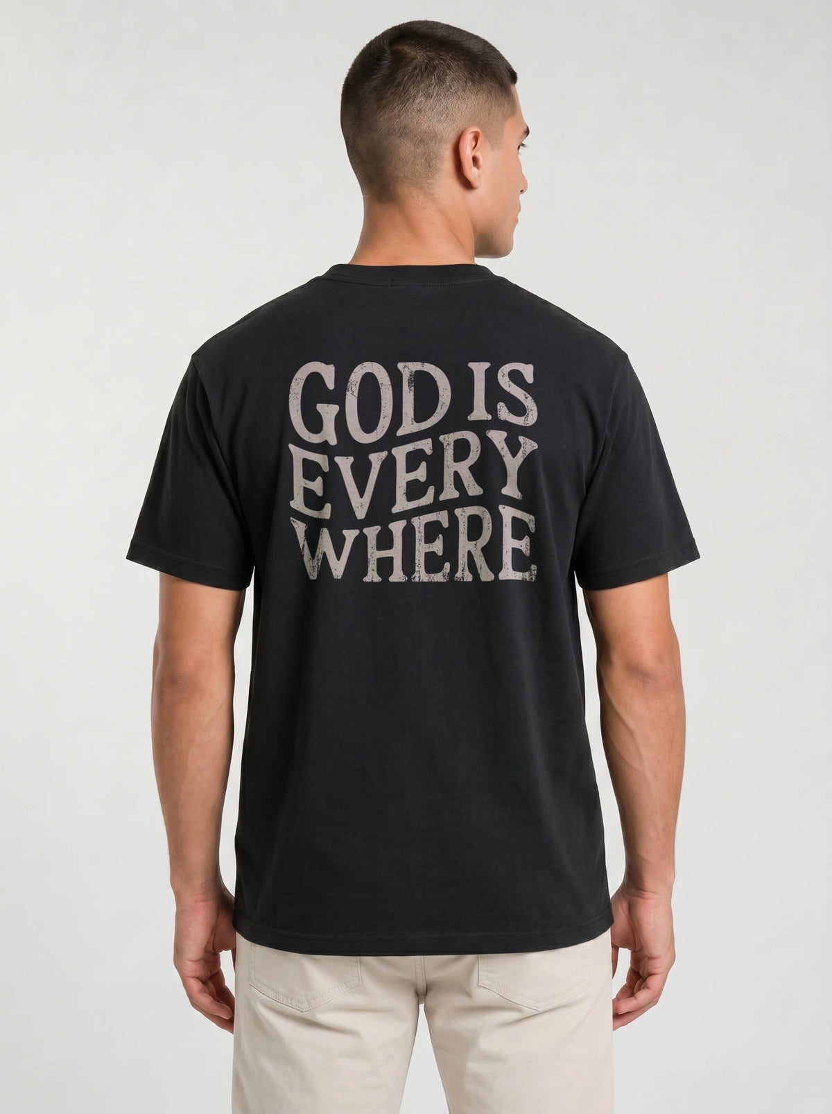 A man viewed from the back wearing a black t-shirt with the phrase 'GOD IS EVERY WHERE' printed in distressed white capital letters across the back, paired with light beige pants, against a light gray studio background.