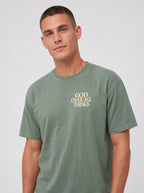 Person wearing a washed sage green T-shirt with a small chest graphic reading “God Over All Things.”