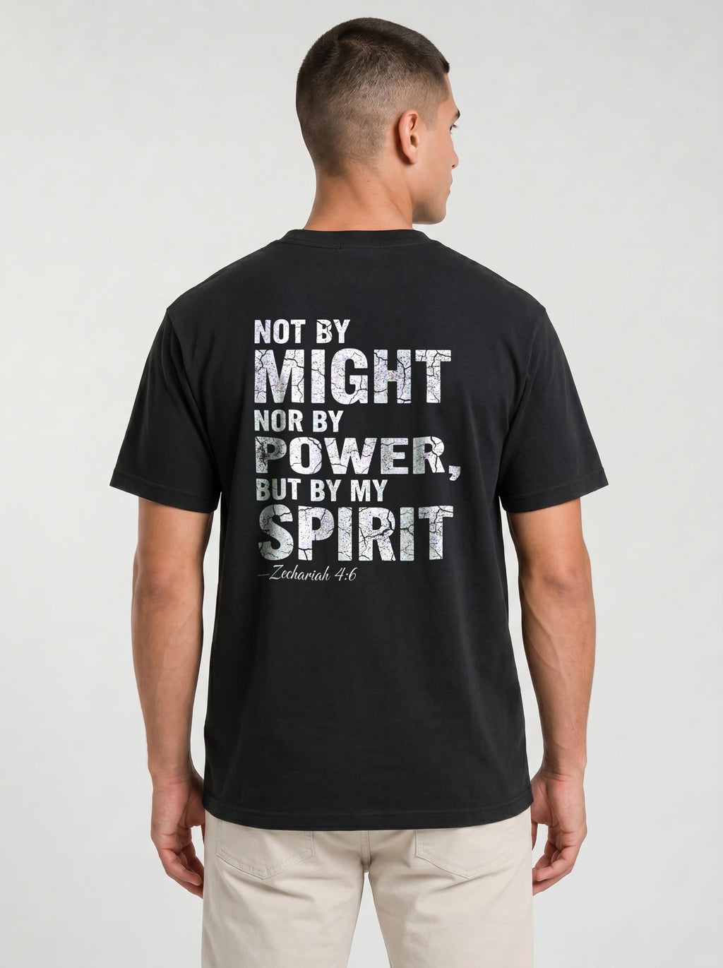 A man viewed from the back wearing a black t-shirt with a distressed white typographic print that reads 'NOT BY MIGHT NOR BY POWER, BUT BY MY SPIRIT -Zechariah 4:6' underneath. He is also wearing light beige pants, set against a plain light gray studio background.