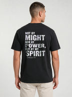 A man viewed from the back wearing a black t-shirt with a distressed white typographic print that reads 'NOT BY MIGHT NOR BY POWER, BUT BY MY SPIRIT -Zechariah 4:6' underneath. He is also wearing light beige pants, set against a plain light gray studio background.