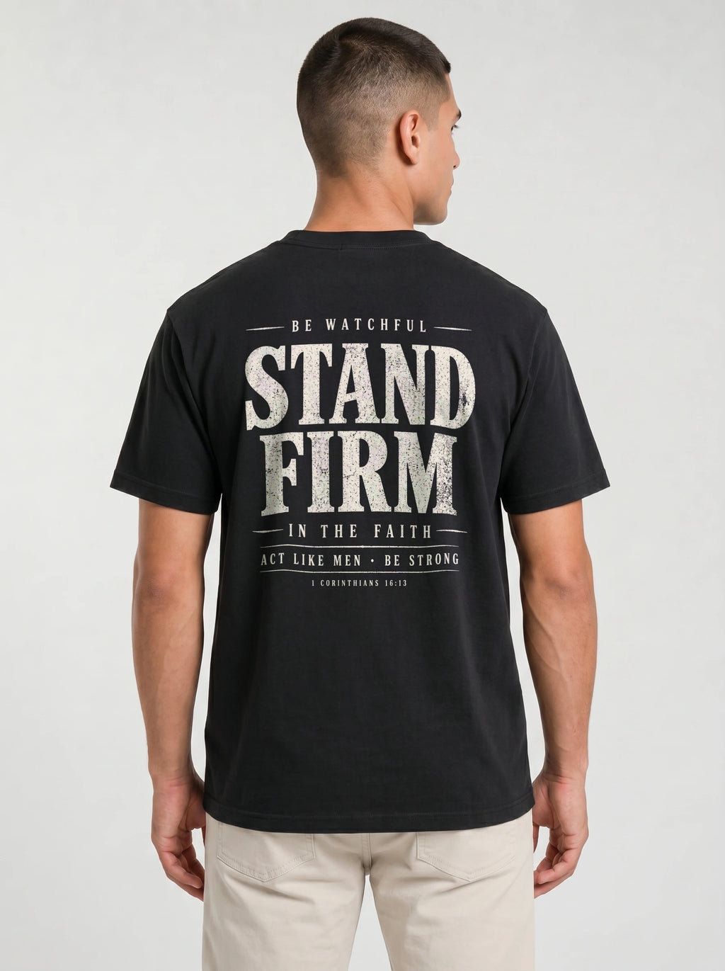A man viewed from the back wearing a black t-shirt with a large distressed white graphic print that reads 'STAND FIRM' and 'BE WATCHFUL IN THE FAITH ACT LIKE MEN - BE STRONG'. He is also wearing light beige pants against a light gray studio background.