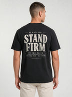 A man viewed from the back wearing a black t-shirt with a large distressed white graphic print that reads 'STAND FIRM' and 'BE WATCHFUL IN THE FAITH ACT LIKE MEN - BE STRONG'. He is also wearing light beige pants against a light gray studio background.