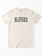 A light beige crewneck t-shirt with the word 'BLESSED' printed across the chest in a dark green collegiate-style font, laid flat against a white background.
