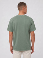 A man viewed from the back wearing a muted sage green short-sleeve t-shirt and light beige pants, standing against a light gray studio background.