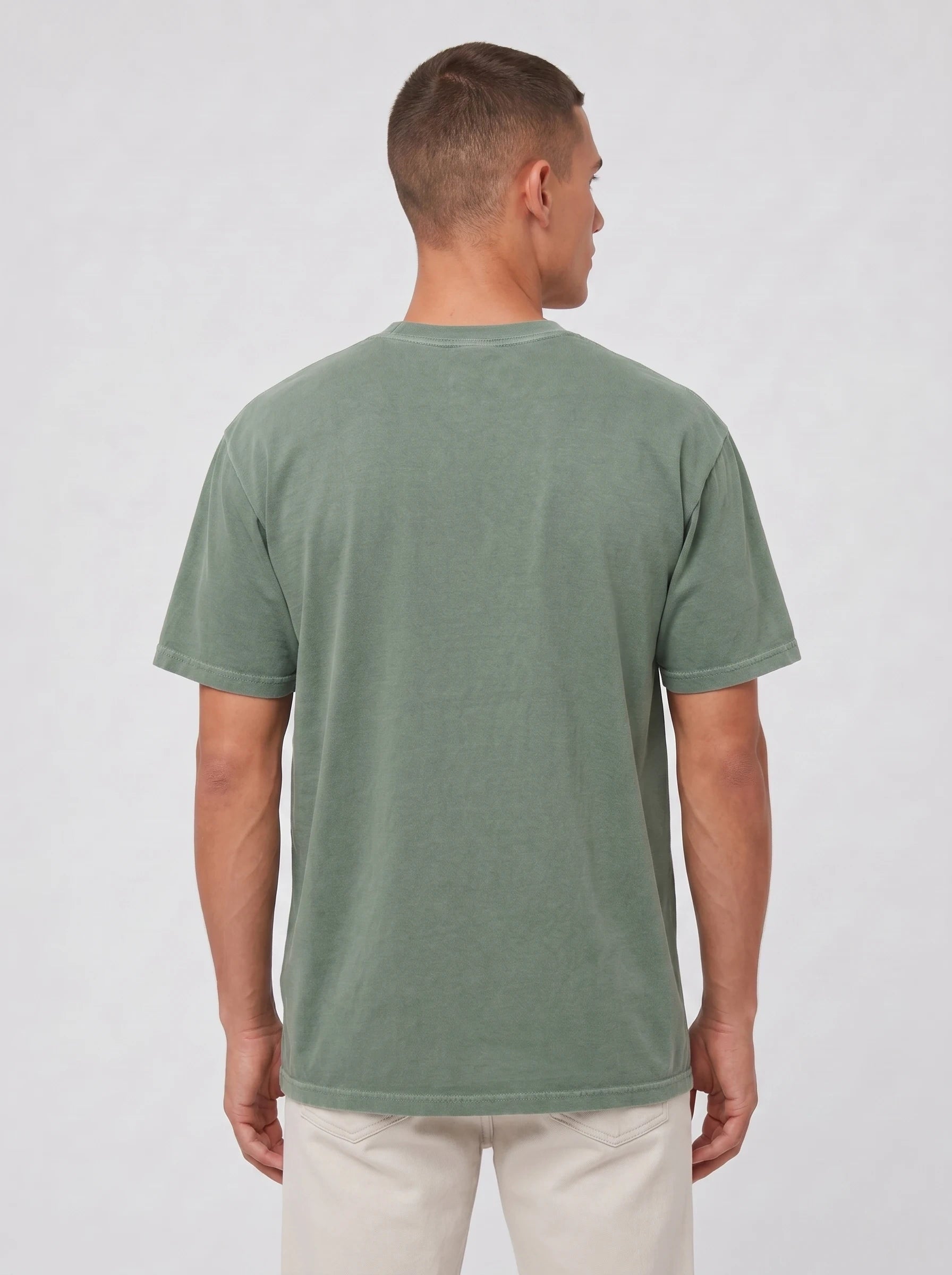 A man viewed from the back wearing a muted sage green short-sleeve t-shirt and light beige pants, standing against a light gray studio background.