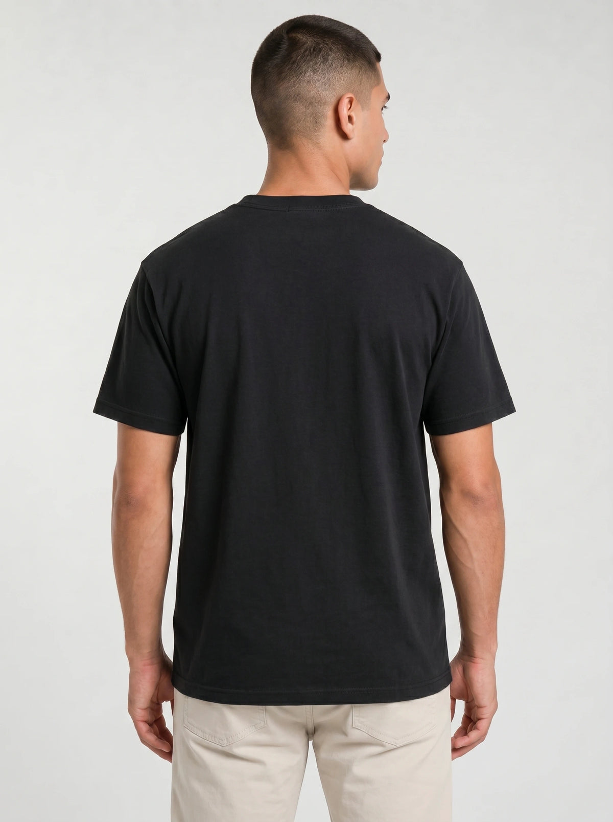 Back view of a man wearing a plain black Comfort Colors t-shirt with no print, paired with beige pants, against a plain light gray studio background.