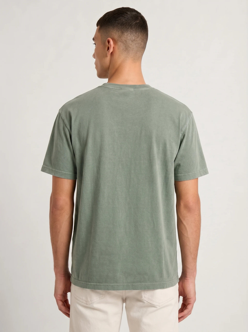 A man viewed from the back wearing a muted sage green short-sleeve t-shirt and off-white trousers, standing against a light gray studio background.