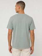 A man viewed from the back wearing a light sage green crewneck t-shirt and off-white trousers, standing against a neutral light gray studio background.