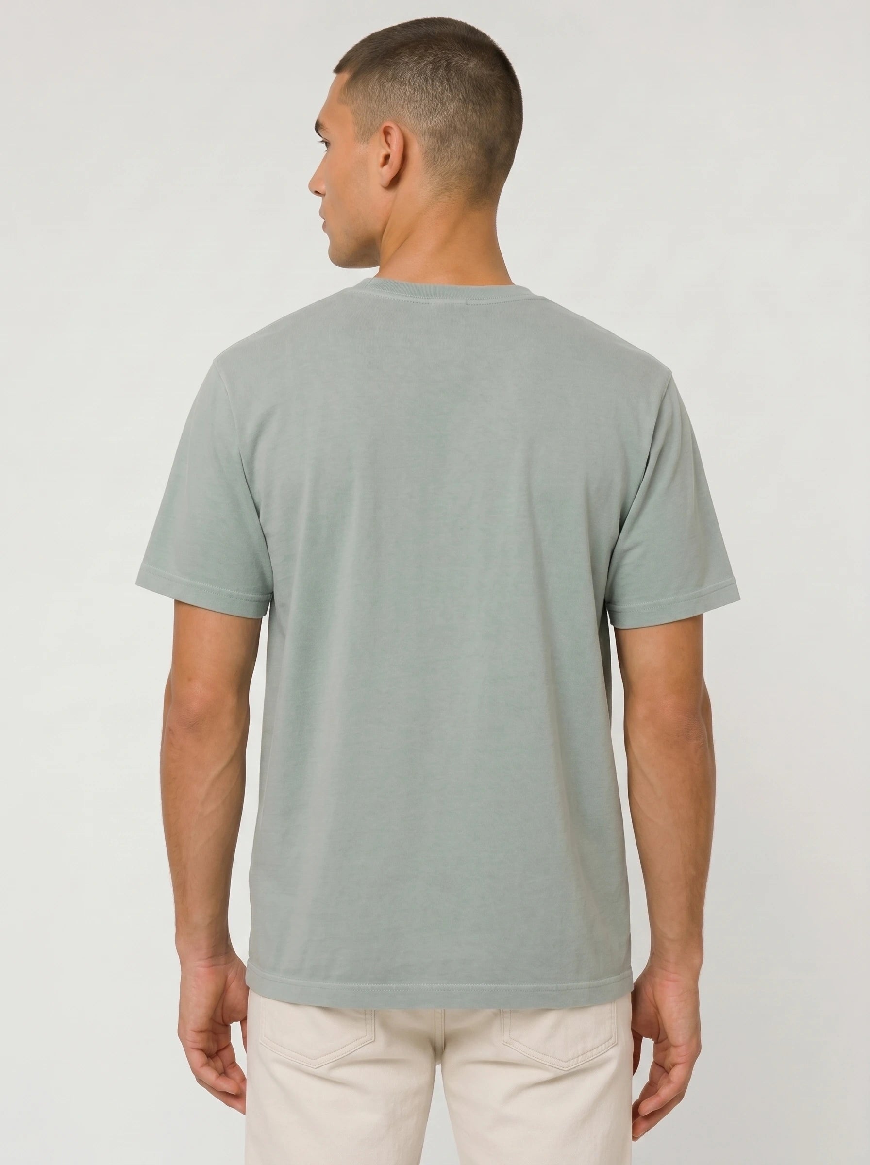 A man viewed from the back wearing a light sage green crewneck t-shirt and off-white trousers, standing against a neutral light gray studio background.
