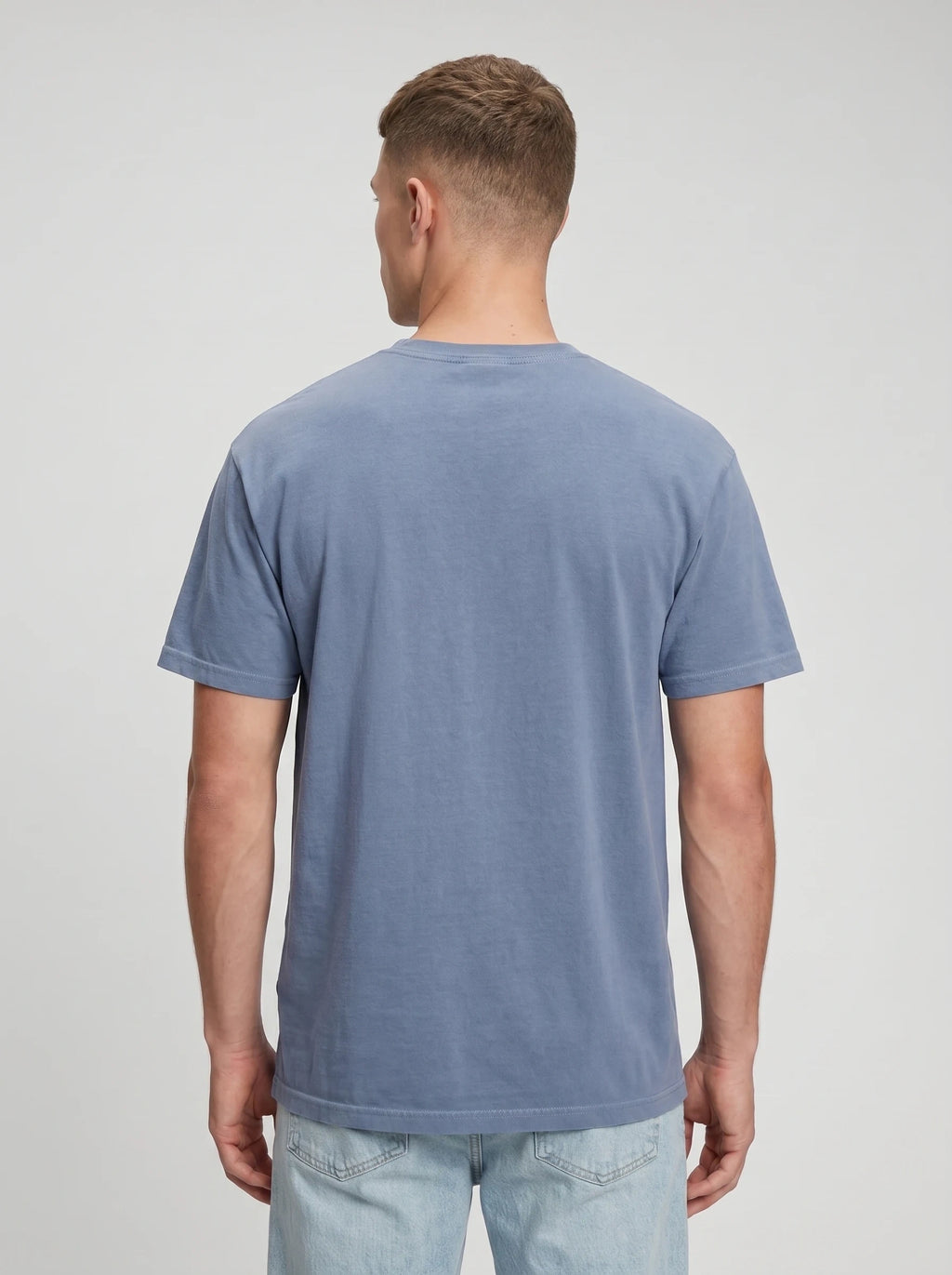 A man viewed from the back wearing a muted slate blue crewneck t-shirt and light wash jeans, set against a plain light gray studio background.