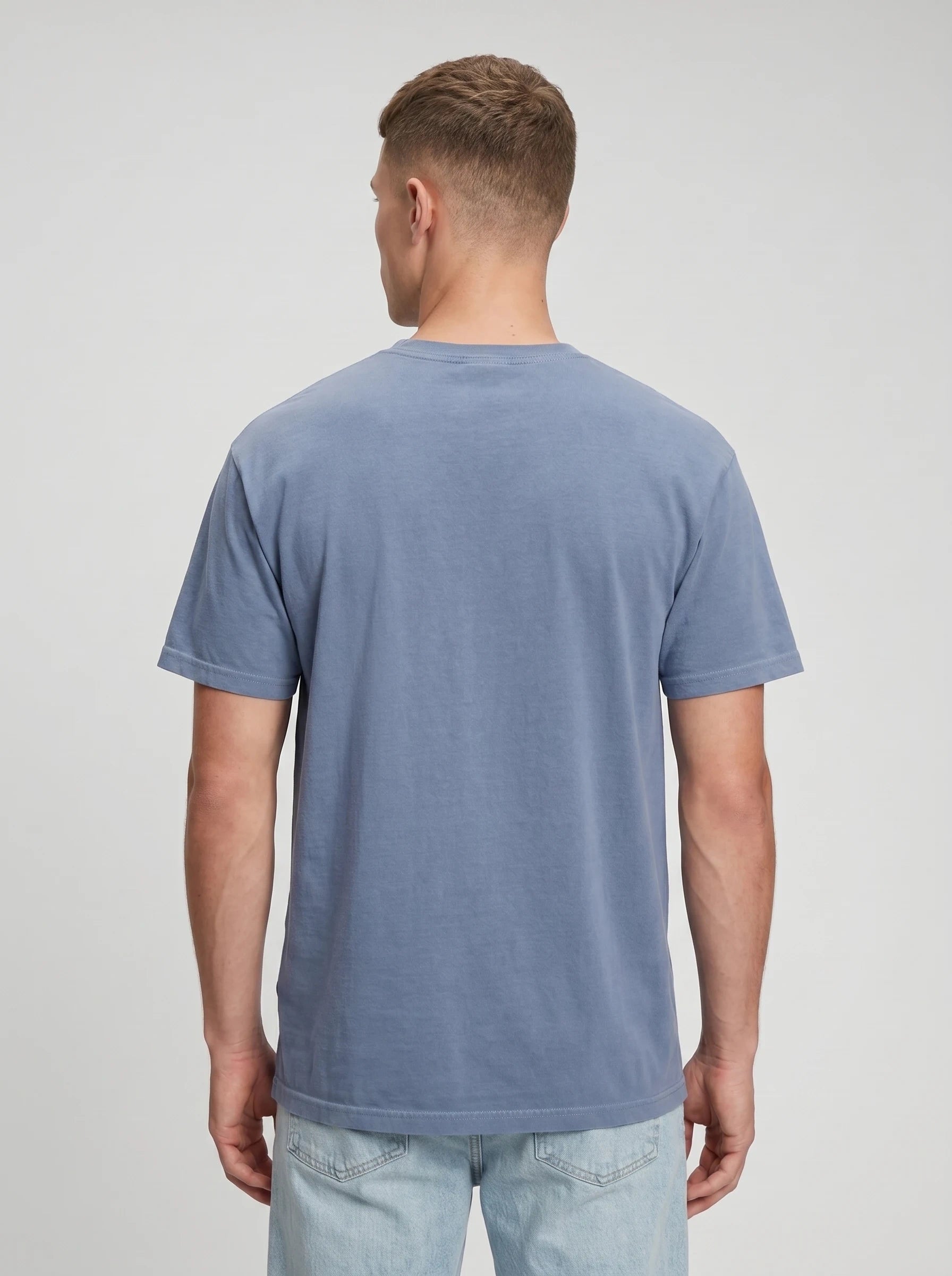 A man viewed from the back wearing a muted slate blue crewneck t-shirt and light wash jeans, set against a plain light gray studio background.