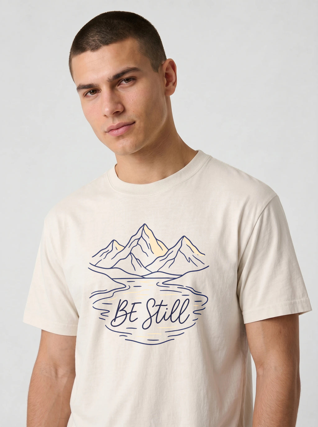 A man wearing a cream-colored t-shirt with a graphic of mountains, a lake, and the text 'BE Still' in navy blue and yellow.