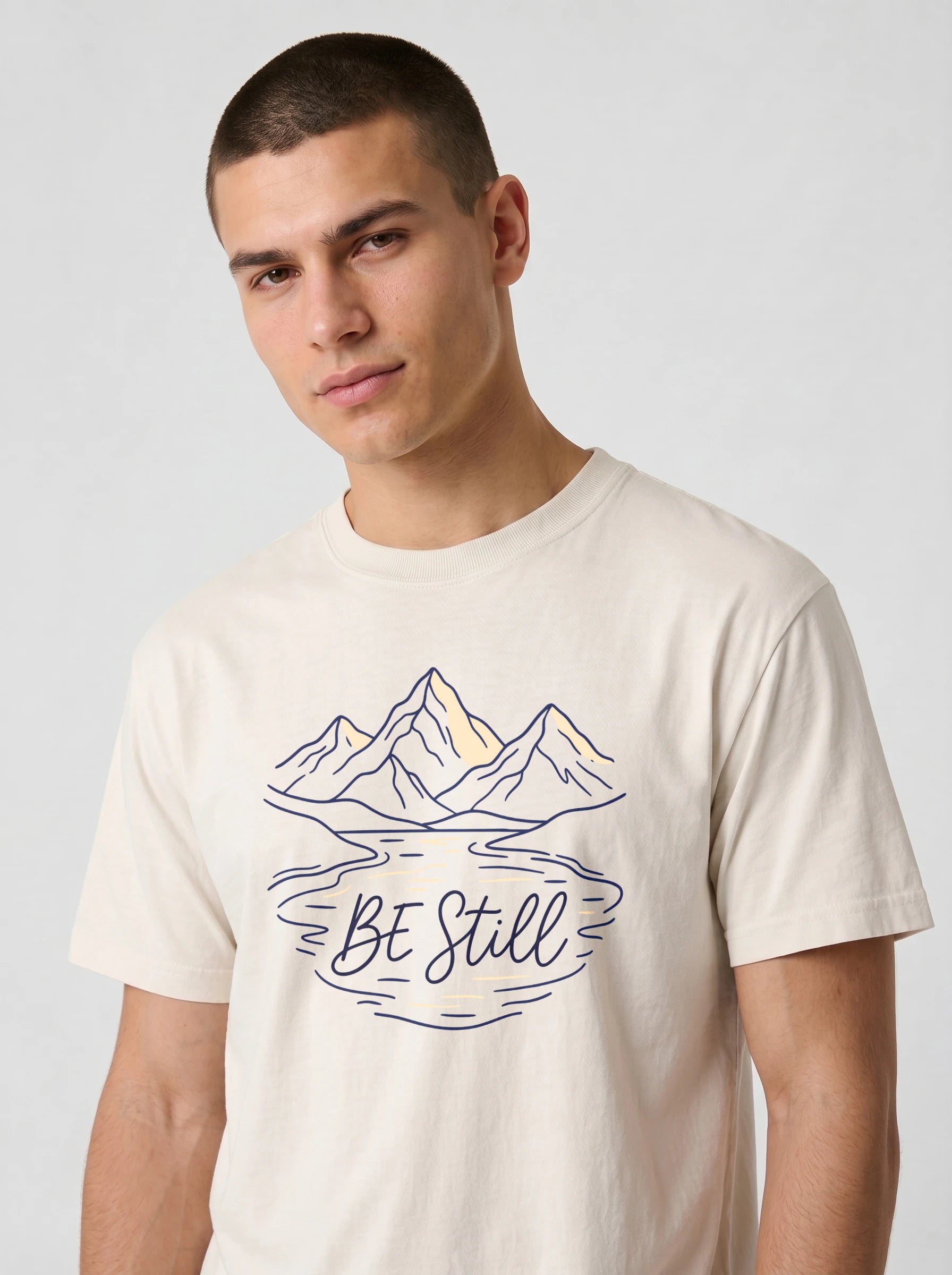 A man wearing a cream-colored t-shirt with a graphic of mountains, a lake, and the text 'BE Still' in navy blue and yellow.