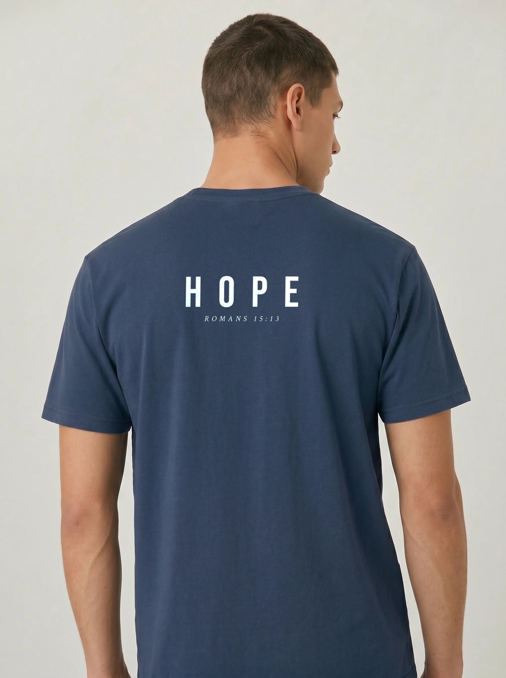 A man viewed from the back wearing a navy blue t-shirt with 'HOPE' printed in white block letters with 'ROMANS 15:13' underneath printed in white on the back, set against a plain light gray background.