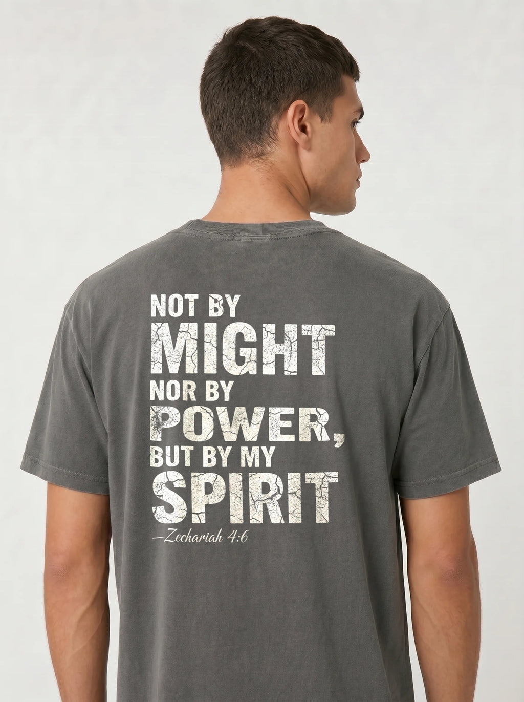 A man viewed from the back wearing a dark gray t-shirt with a distressed white typographic print that reads 'NOT BY MIGHT NOR BY POWER, BUT BY MY SPIRIT - Zechariah 4:6'.