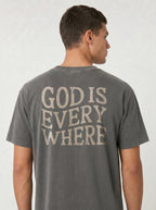A man viewed from the back wearing a dark gray t-shirt with the phrase 'GOD IS EVERY WHERE' printed in a distressed cream-colored font across the back.
