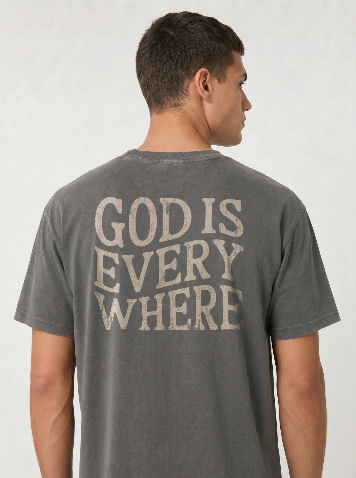 A man viewed from the back wearing a dark gray t-shirt with the phrase 'GOD IS EVERY WHERE' printed in a distressed cream-colored font across the back.