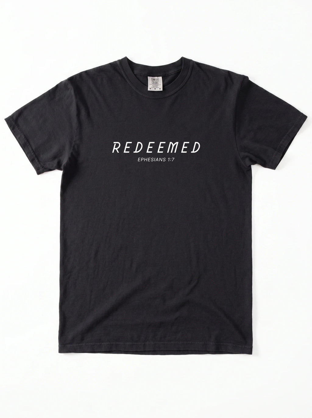 Flat lay of a folded black Comfort Colors t-shirt printed with "REDEEMED" in bold white capital letters and "EPHESIANS 1:7" underneath, on a plain white background.