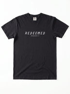 Flat lay of a folded black Comfort Colors t-shirt printed with "REDEEMED" in bold white capital letters and "EPHESIANS 1:7" underneath, on a plain white background.