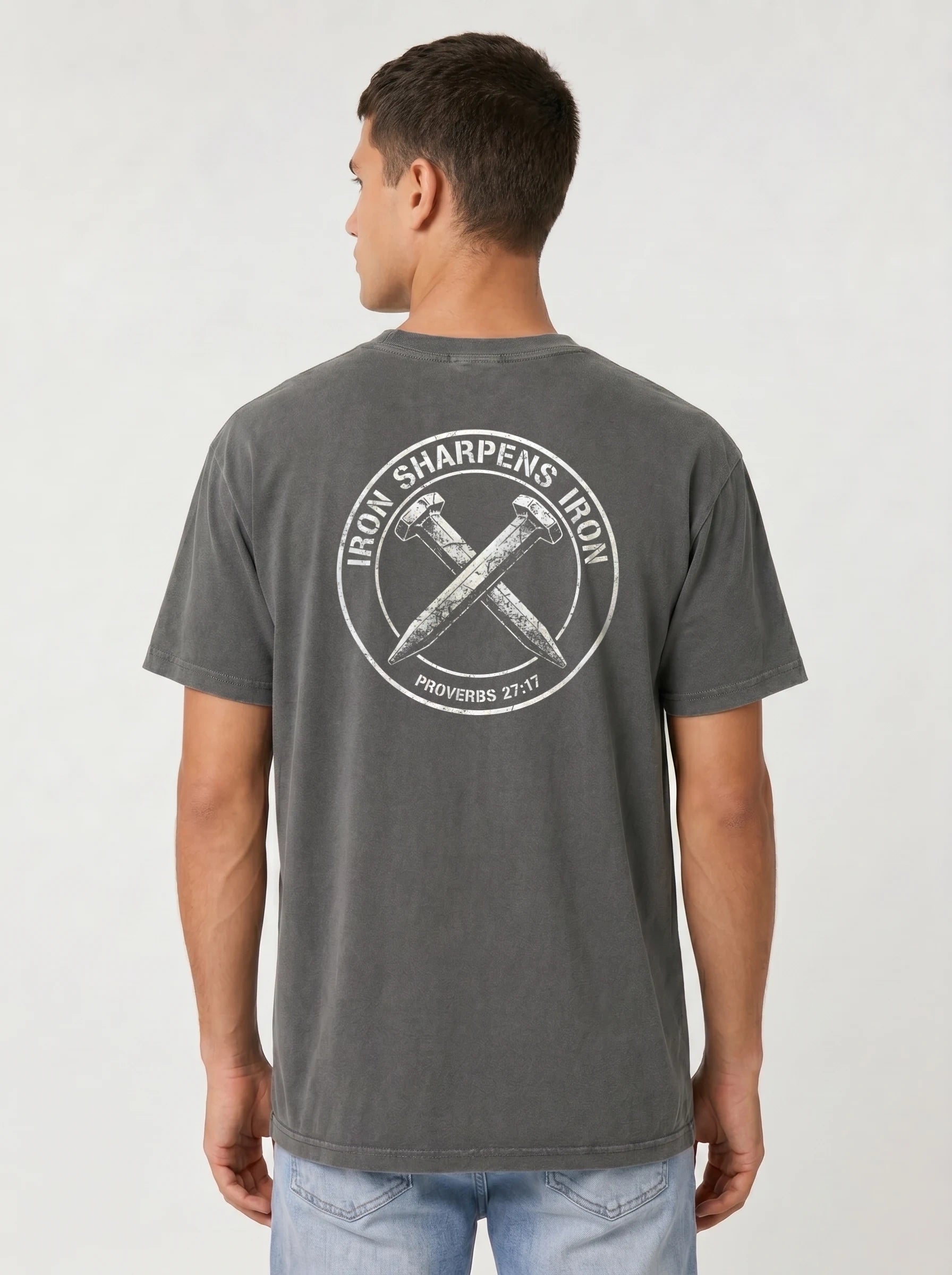 A man viewed from the back wearing a dark gray t-shirt with a large white circular graphic on the back featuring crossed hammers and the text 'IRON SHARPENS IRON' and 'PROVERBS 27:17'.