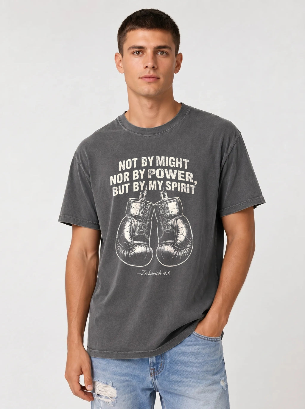 A man wearing a dark gray graphic t-shirt with a white boxing theme and the quote 'NOT BY MIGHT NOR BY POWER, BUT BY MY SPIRIT' paired with light blue jeans, against a light gray studio background.
