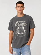 A man wearing a dark gray graphic t-shirt with a white boxing theme and the quote 'NOT BY MIGHT NOR BY POWER, BUT BY MY SPIRIT' paired with light blue jeans, against a light gray studio background.