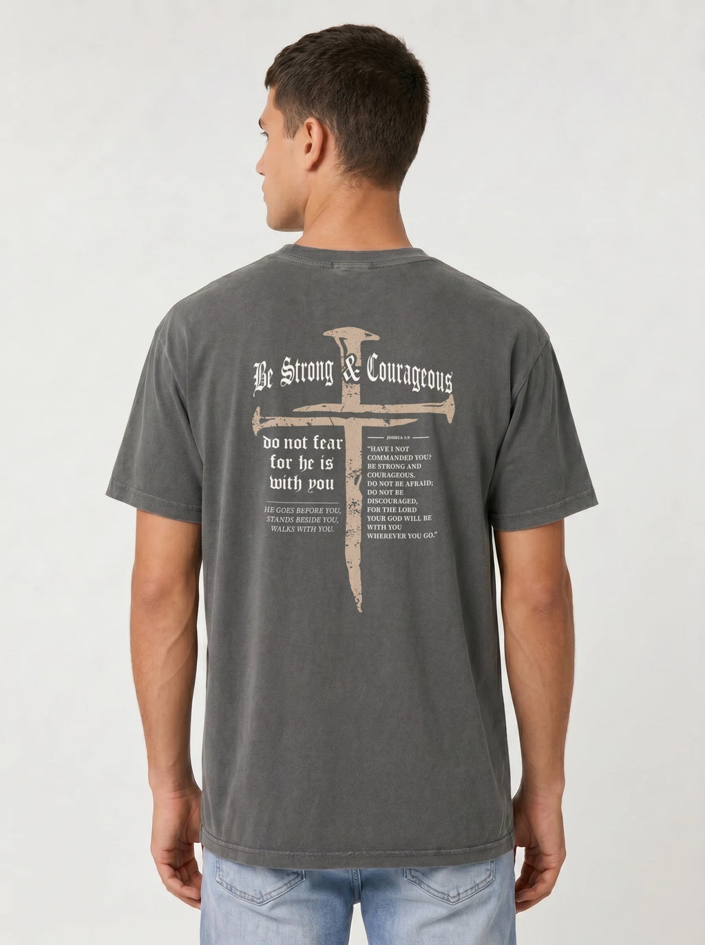 A man wearing a dark gray washed t-shirt from the back, featuring a large graphic print of a cross with a nail through it and the text 'Be Strong & Courageous' and 'do not fear for he is with you'.